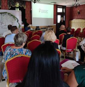 Pokaż zdjęcie: Grupa uczestników siedzi na czerwonych krzesłach, słuchając prezentacji na temat projektów realizowanych przez Państwowy Fundusz Rehabilitacji Osób Niepełnosprawnych. Na ekranie wyświetlone jest logo PFRON. Po prawej stronie ekranu stoi kobieta w czarnym żakiecie a po lewej przy stoliku siedzi kobieta w okularach. 