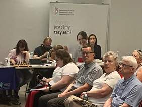 Pokaż zdjęcie: Grupa osób siedzi w sali konferencyjnej. Z boku po prawej kobieta w ciemnych włosach. W tle baner z logo PFRON  i z napisem „Wszyscy jesteśmy tacy sami”.