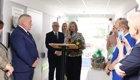 Pokaż zdjęcie: Grupa osób bierze udział w ceremonii otwarcia nowego pomieszczenia w szpitalu. Na pierwszym planie widoczna jest kobieta, która przemawia do zgromadzonych, trzymając bukiet kwiatów. Obok niej stoi mężczyzna w garniturze, a w tle widać inne osoby oraz plakat informacyjny o szpitalu.