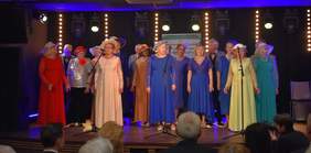 Pokaż zdjęcie: Grupa seniorów występuje na scenie podczas obchodów Światowego Dnia Inwalidy w Białymstoku. Kobiety ubrane są w długie, eleganckie suknie w różnych kolorach oraz kapelusze, a mężczyźni w garnitury i muszki. W tle widoczne są reflektory sceniczne oraz baner Regionalnego Ośrodka Polityki Społecznej w Białymstoku. Przed sceną siedzi publiczność, a fotograf uwiecznia występ.