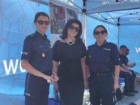 Pokaż zdjęcie: Trzy kobiety stojące razem i uśmiechające się do obiektywu. Dwie z nich, po lewej i prawej stronie, ubrane są w granatowe mundury policyjne z widocznym napisem „POLICJA” oraz naszywkami i identyfikatorami. Kobieta pośrodku – dyrektor Oddziału ma na sobie elegancką, czarną sukienkę. Wszystkie trzy mają ciemne okulary przeciwsłoneczne. Zdjęcie wykonano na tle niebieskich namiotów z napisem podczas wydarzenia plenerowego.