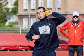 Pokaż zdjęcie: Mężczyzna w ciemnym dresie i okularach trzyma przy szyi kulę. Jest w pozycji przygotowanej do rzutu.  Na klace ma nr 40 nad nim Logo PFRON.