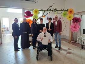 Pokaż zdjęcie: Grupa siedmiu osób w jasnej sali z oknami. Na pierwszym planie uśmiechnięty mężczyzna na wózku i trzyma torbę z prezentem. Za nim stoi sześć osób. W tle ściana ozdobiona dużymi, kolorowymi kwiatami z papieru oraz stoliki w dalszej części sali.