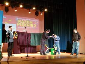 na scenie osoba próbuje swoich sił jako aktor lalkarz, aktor mu pomaga, obok sto mężczyzna, uśmiecha się w lewym rogu dwie osoby konferansjerów,