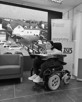 Pokaż zdjęcie: Dr Anna Drabarz, kobieta siedząca na wózku elektrycznym, prowadzi panel dyskusyjny podczas XIII Konferencji „Dostępność dziś i jutro” w Białymstoku. Ubrana w jasny sweter i granatowe spodnie, spogląda z zaangażowaniem w stronę uczestników. W tle widoczny jest ekran z obrazem budynku oraz bannery instytucji wspierających wydarzenie, w tym PFRON i ZUS.