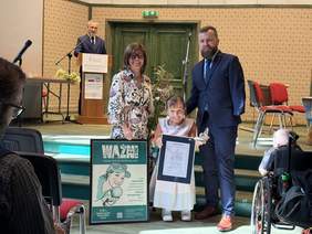 Pokaż zdjęcie: Na scenie stoją trzy osoby – kobieta w kolorowej sukience, mężczyzna w garniturze oraz uśmiechnięta kobieta w jasnej sukni trzymająca w rękach certyfikat i statuetkę motyla. Obok nich na podłodze stoi duży plakat z napisem „WAŻNI”. W tle widać prowadzącego przy mównicy oraz czerwone krzesła.