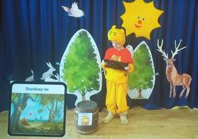 Pokaż zdjęcie: Scena na której dziecko przebrane za Kubusia Puchatka korzysta z urządzenia wspomagania mowy, w tle są kolorowe dekoracje takie jak drzewa, jelonki, słońce