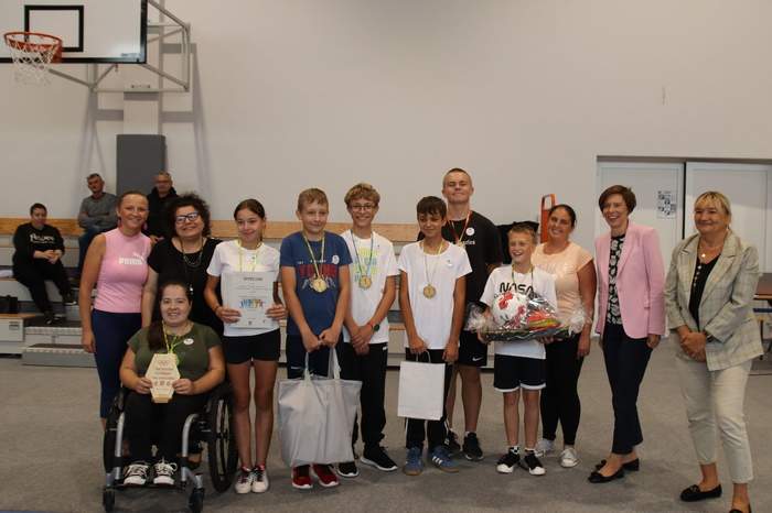 Pokaż zdjęcie: Grupa kilkunastu uśmiechniętych osób stoi w rzędzie wewnątrz hali sportowej. Część osób ma na szyi medale, a niektóre osoby trzymają także dyplomy oraz nagrody rzeczowe. W tle możemy dostrzec kosz do gry w koszykówkę.