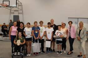 Pokaż zdjęcie: Grupa kilkunastu uśmiechniętych osób stoi w rzędzie wewnątrz hali sportowej. Część osób ma na szyi medale, a niektóre osoby trzymają także dyplomy oraz nagrody rzeczowe. W tle możemy dostrzec kosz do gry w koszykówkę.