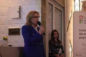 Pokaż zdjęcie: Kobieta blondynka w niebieskim garniturze stoi z mikrofonem i wita uczestników konferencji, na drugim planie kobieta z długimi włosami trzyma w ręku dokumenty i spogląda na kobietę przemawiającą przez mikrofon