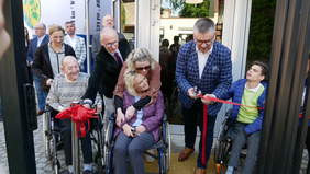 Pokaż zdjęcie: Mieszkańcy Powiatu Strzeleckiego - osoby starsze i osoby z niepełnosprawnościami przecinają wstęgę uroczyście otwierając windę przy budynku Starostwa Powiatowego