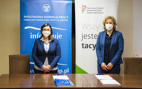 Pokaż zdjęcie: dwie kobiety w niebieskich marynarkach stoją przy brązowym stole, w tle loga PIP oraz PFRON