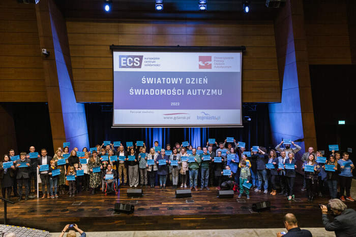 Pokaż zdjęcie: Na scenie stoi wiele ludzi trzymających niebieskie kartki z napisem w rękach. Nad nimi wisi wielki ekran na którym jest napis Światowy Dzień Świadomości Autyzmu 2023