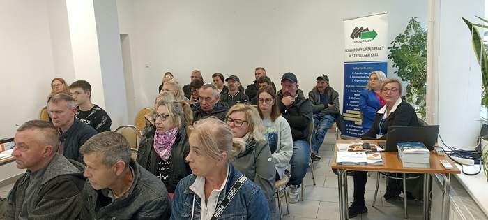 Pokaż zdjęcie: Uczestnicy spotkania siedzą na krzesłach w oświetlonej sali