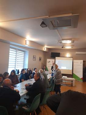 Pokaż zdjęcie: Sala w której przy stole siedzi grupa osób, z przodu sali stoi ekran oraz rollupy z logo PFRON oraz OWiT.