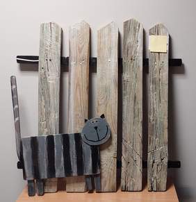 Pokaż zdjęcie: drewniana rzeźba przedstawia szarego w czarne paski kota a za nim płot