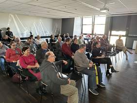 Pokaż zdjęcie: Na Sali siedzą w rzędach ludzie. Patrzą się w jedną stronę. Za nimi rysunki dźwigów stoczni gdańskiej. Po prawej duże okna.