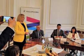 Pokaż zdjęcie: Kobieta stoi na środku i przemawia. Przy stole siedzi dwóch mężczyzn i kobieta. Wszyscy są elegancko ubrani.