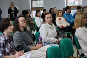 Pokaż zdjęcie: Grupa kilkunastu osób znajduje się w sali szkoleniowej i siedzi na zielonych krzesłach. Część z tych osób trzyma w dłoniach kubek z napojem, długopis lub notatki. Z tyłu sali stoi ponadto jeszcze jedna kobieta.