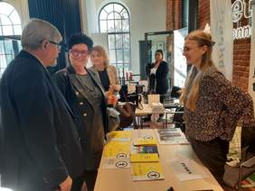 Pokaż zdjęcie: Przy stoisku informacyjnym PFRON stoją dwie kobiety i jeden mężczyzna. Panie uśmiechają się. W tle widać inne osoby. Na stole leżą różne informatory, broszury, ulotki i materiały reklamowe. 