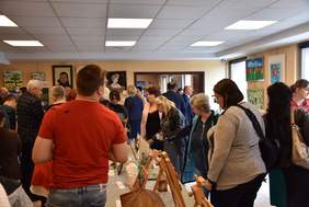 Pokaż zdjęcie: W dużej sali na ścianach są zawieszone obrazy, na środku stoją stoły z ustawionymi na nich rzeźbami. Kilkanaście osób chodzi i ogląda wystawione prace.