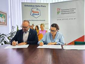 Pokaż zdjęcie: Przedstawiciele Lubelskiej Wojewódzkiej Komendy OHP i Oddziału Lubelskiego PFRON podpisują porozumienie o współpracy przy stole, na tle banerów obu instytucji.