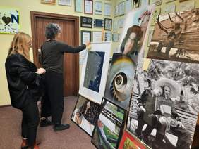 Pokaż zdjęcie: Na sztalugach wiszą przeróżne fotografie. Przed nimi stoją dwie kobiety, które z uwagą przyglądają się zdjęciom. 