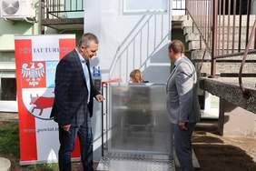 Pokaż zdjęcie: Prezentacja nowego podnośnika dla osób z niepełnosprawnościami przy zewnętrznych schodach budynku. Dwóch elegancko ubranych mężczyzn stoi po obu stronach przeszklonej, pionowej platformy windy, w której widać siedzącą osobę. W tle widoczne są betonowe schody z metalową barierką prowadzące w górę.