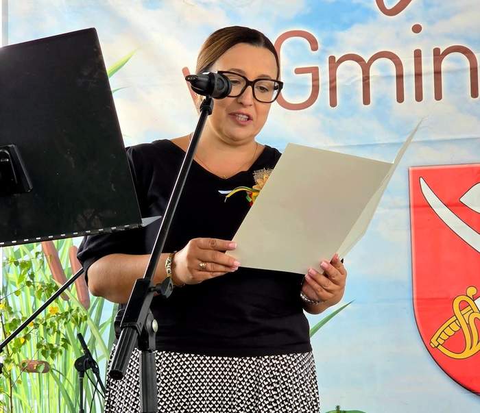 Pokaż zdjęcie: Kobieta w okularach czyta list podczas dożynek. Ubrana jest w czarną bluzkę i czarno-białą spódnicę.