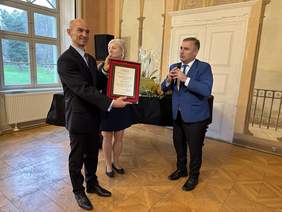 Pokaż zdjęcie: Mężczyzna w czarnych spodniach, niebieskiej marynarce i krawacie trzyma w rękach mikrofon. Obok niego mężczyzna w ciemnym garniturze trzyma w rękach grawer i się uśmiecha. Za nim kobieta w blond włosach patrzy w stronę widowni również się uś