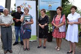 Pokaż zdjęcie: Dwóch mężczyzna i cztery kobiety stoją na chodniku. W tle widoczna jest tabliczka z logotypem Środowiskowego Domu Samopomocy w Doruchowie. Jedna z kobiet przemawia do mikrofonu.
