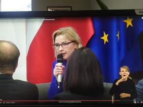 Pokaż zdjęcie: Na tle flagi Polski i Unii Europejskiej siedzi kobieta z mikrofonem w niebieskim garniturze, lekko przysłonięta głowami osób siedzących na widowni. W prawym dolnym rogu zdjęcia widać kobiecą postać tłumacza języka migowego