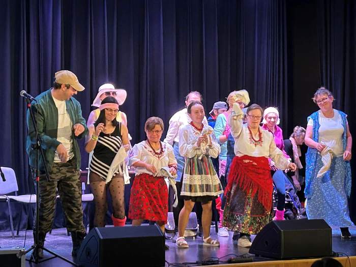 Pokaż zdjęcie: Grupa kilkunastu osób, głównie kobiet, stoi na scenie teatralnej i jest ubrana w wielobarwne stroje. Na scenie ustawiony jest mikrofon, jak również znajdują się dwa odsłuchy oraz kilka krzeseł.