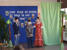 Pokaż zdjęcie: na zdjęciu hol szkoły, na której umieszczono scenografię do sztuki, trójka dzieci ma na głowie maski z postacią słonia z trąbą do góry, przebrana w postacie ktróre gra dla publiczności