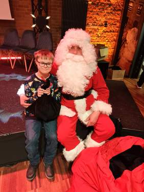 Pokaż zdjęcie: Obok Świętego Mikołaja stoi chłopiec z Zespołem Downa. W dłoniach trzyma prezent. 