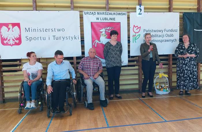 Pokaż zdjęcie: Sala gimnastyczna, przy drabinkach, po lewej stronie siedzą trzy osoby na wózkach inwalidzkich, po prawej stronie stoją trzy kobiety. Jedna z kobiet trzyma w ręku mikrofon.