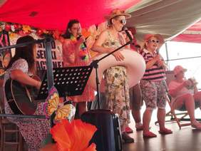 Pokaż zdjęcie: Na scenie znajduje sie zespół śpiewający piosenkę. Dwie dziewczyny śpiewają do mikrofonu, jedna gra na gitarze klasycznej, a jeszcze inna siedzi na leżaku i udaje, że pije drinka. 