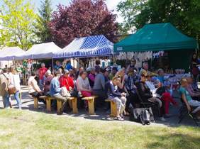 Pokaż zdjęcie: miejsce w plenerze rozstawione ławki na których siedzą zaproszeni goście  w tle stoiska z wyrobami WTZ