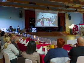 Pokaż zdjęcie: Sala udekorowana na jubileusz, ustawione stoły z gośćmi, na scenie duży ekran z projekcją zdjęć, obok świecące dekoracje z liczbą 25 i skrzydłami. Atmosfera uroczystej gali lub rocznicy.