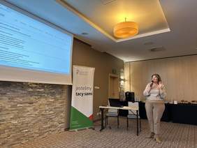 Pokaż zdjęcie: Sala konferencyjna. Po lewej stronie wisi ekran do rzutnika z prezentacją z napisem Wróć. Obok baner z logo PFRON z napisem wszyscy jesteśmy tacy sami. Przy nim stolik z laptopami. Obok stoi kobieta w okularach ubrana w jasny sweter i jasne spodnie. T