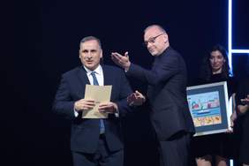 Pokaż zdjęcie: Mężczyzna w Granatowym garniturze, białej koszuli i krawacie trzyma w ręku dokument z napisem Gdynia, obok niego mężczyzna w ciemnym garniturze, krótko przyciętych włosach i okularach wskazuje na coś ręką, w tle kobieta w ciemnej sukience trzy