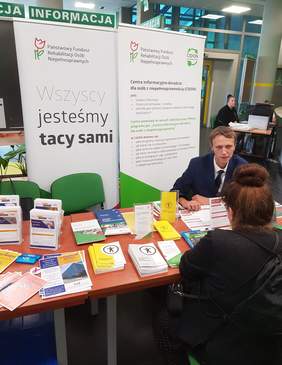 Pokaż zdjęcie: Mężczyzna siedzi za stolikiem na tle baneru PFRON oraz CIDON i udziela informacji kobiecie, która przyszła do ZUS na Dzień Osób z Niepełnosprawnością. Na stoliku ułożone są materiały informacyjne promujące programy i zadania PFRON.