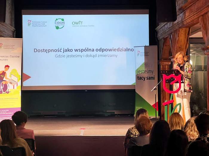 Pokaż zdjęcie: Na scenie, przed tulipanem z logo PFRON wykonanym z tworzywa sztucznego, stoi kobieta, która przemawia do osób zgromadzonych na sali. Na dużym ekranie, pomiędzy banerami organizatorów konferencji wyświetlony jest slajd z tytułem prelekcji.