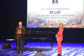 Pokaż zdjęcie: Mężczyzna w ciemnym garniturze stoi na scenie po lewej stronie, trzymając statuetkę w kształcie serca, podczas gdy kobieta w różowym garniturze stoi obok niego po prawej. Obaj są oświetleni niebieskim światłem. Za nimi znajduje się duży ekran z napisem po polsku: "Koło PSONI W STARGARDZIE 25 LAT ZAKŁADU AKTYWNOŚCI ZAWODOWEJ "CENTRALNA KUCHNIA"". Na ekranie w tle widać też zdjęcia przygotowanych potraw. Na pierwszym planie, na podłodze, stio czarny fortepian.