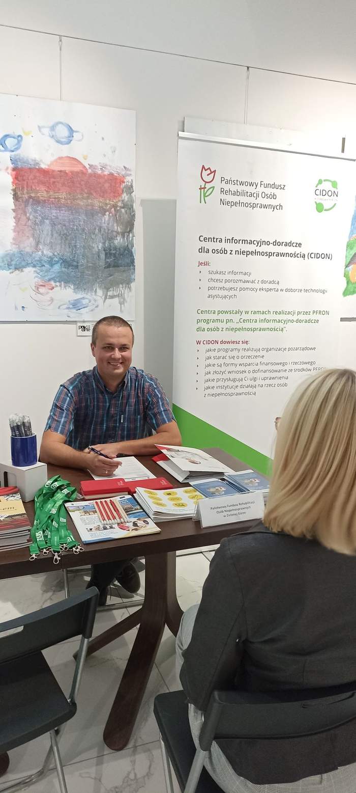 Pokaż zdjęcie: Przedstawiciel Oddziału Lubuskiego PFRON przy stoliku udziela informacji zainteresowanej osobie, jest uśmiechnięty w koszule w kratkę, na tle roll-up z logo CIDON