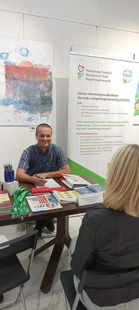 Pokaż zdjęcie: Przedstawiciel Oddziału Lubuskiego PFRON przy stoliku udziela informacji zainteresowanej osobie, jest uśmiechnięty w koszule w kratkę, na tle roll-up z logo CIDON