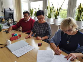 Pokaż zdjęcie: Trzy osoby, mężczyzna i dwie panie siedzą przy jasnym dużym stole nad dokumentami, za nimi ściana okien