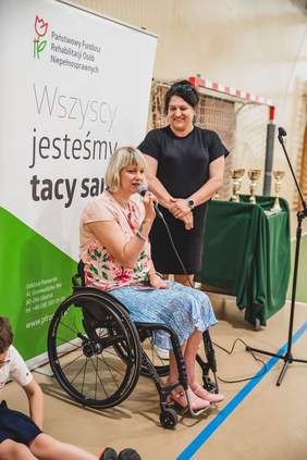 Pokaż zdjęcie: Kobieta w blond włosach siedzi na wózku i mówi do mikrofonu który trzyma w ręku. Obok niej stoi kobieta ubrana na czarno i uśmiecha się patrząc na nią. Za niemi baner z logo Pfron i napisem wszyscy jesteśmy tacy sami.