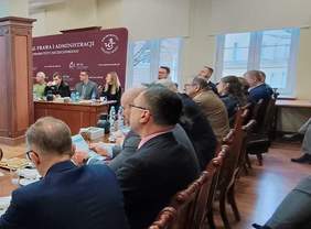 Pokaż zdjęcie: Sala konferencyjna z długim stołem, przy którym siedzi kilkanaście osób w garniturach. Uczestnicy obrad słuchają prelegentów siedzących przy podwyższeniu. Na stołach znajdują się dokumenty, filiżanki, woda i notesy. W tle ekrany prezentacyjne i banery Wydziału Prawa i Administracji.