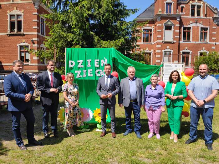 Pokaż zdjęcie: Uczetnicy spotkania stoją obok siebie na tle napisu Dzień Dziecka, na dworze, jest słonecznie